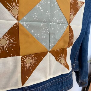 May include: A denim jacket with a patchwork design on the back. The patchwork features a starburst pattern in shades of brown, tan, and light blue. The jacket has a classic collar and button fastenings.