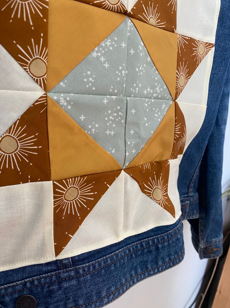 May include: A denim jacket with a patchwork design on the back. The patchwork features a starburst pattern in shades of brown, tan, and light blue. The jacket has a classic collar and button fastenings.