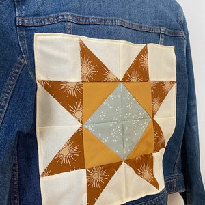 May include: A blue denim jacket with a patchwork star design on the back. The star is made from cream, brown, mustard yellow, and light blue fabrics. The star design includes sunburst details.