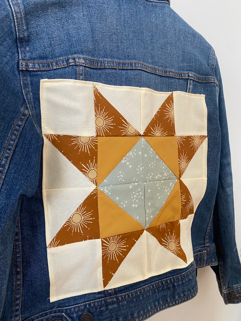 May include: A blue denim jacket with a patchwork star design on the back. The star is made from cream, brown, mustard yellow, and light blue fabrics. The star design includes sunburst details.