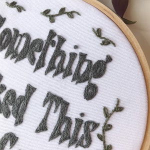 May include: An embroidery hoop with the words "Something good this way comes" stitched in gray thread on white fabric. Small green embroidered leaves and vines are scattered around the text. The hoop is made of natural wood.