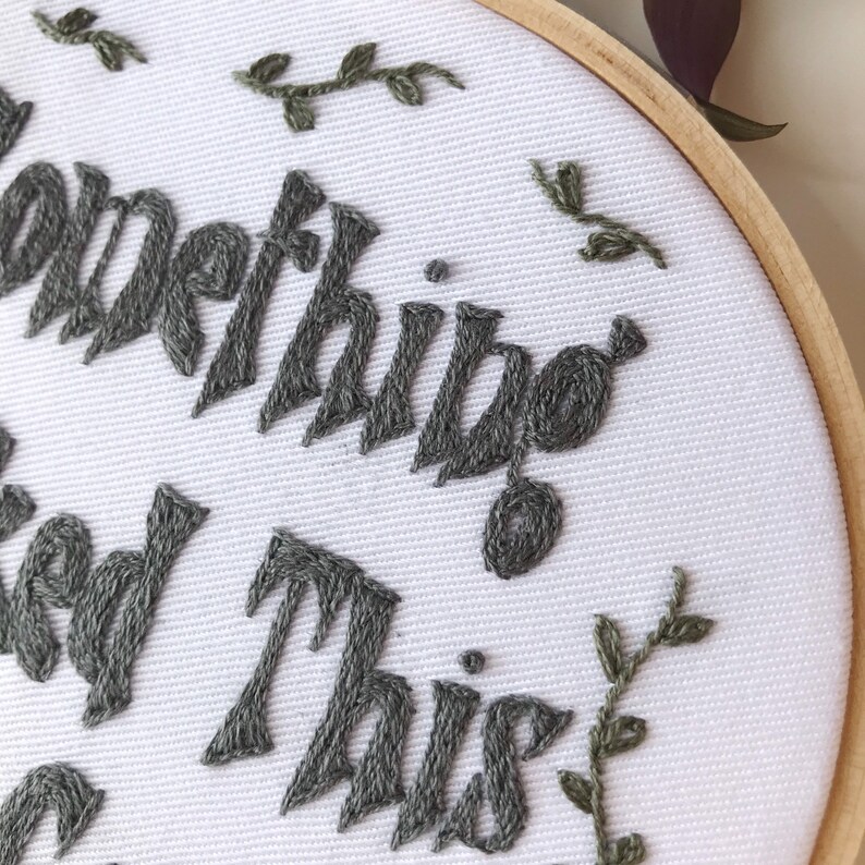 May include: An embroidery hoop with the words "Something good this way comes" stitched in gray thread on white fabric. Small green embroidered leaves and vines are scattered around the text. The hoop is made of natural wood.
