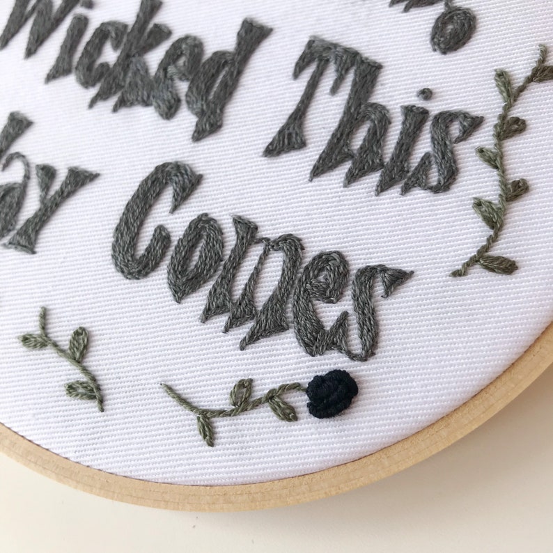 May include: Embroidered hoop art with the phrase "Wicked This Way Comes" in gray thread on white fabric. Features a black rose and green leaf accents. The wooden hoop frame adds a rustic touch to this handmade piece.