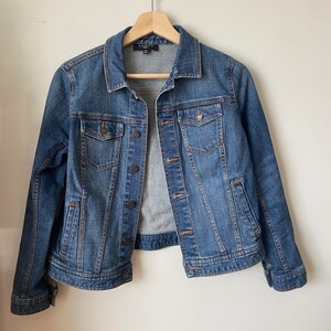May include: A blue denim jacket with a classic design, featuring a collared neckline, button-down front, and multiple pockets. The jacket is hanging on a wooden hanger against a plain white background.