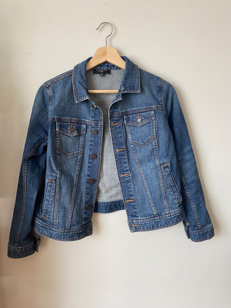 May include: A blue denim jacket with a classic design, featuring a collared neckline, button-down front, and multiple pockets. The jacket is hanging on a wooden hanger against a plain white background.