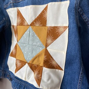 May include: A blue denim jacket with a patchwork star design on the back. The star is made of brown, yellow, and blue fabrics, set against a white background. The star design features a sunburst pattern.