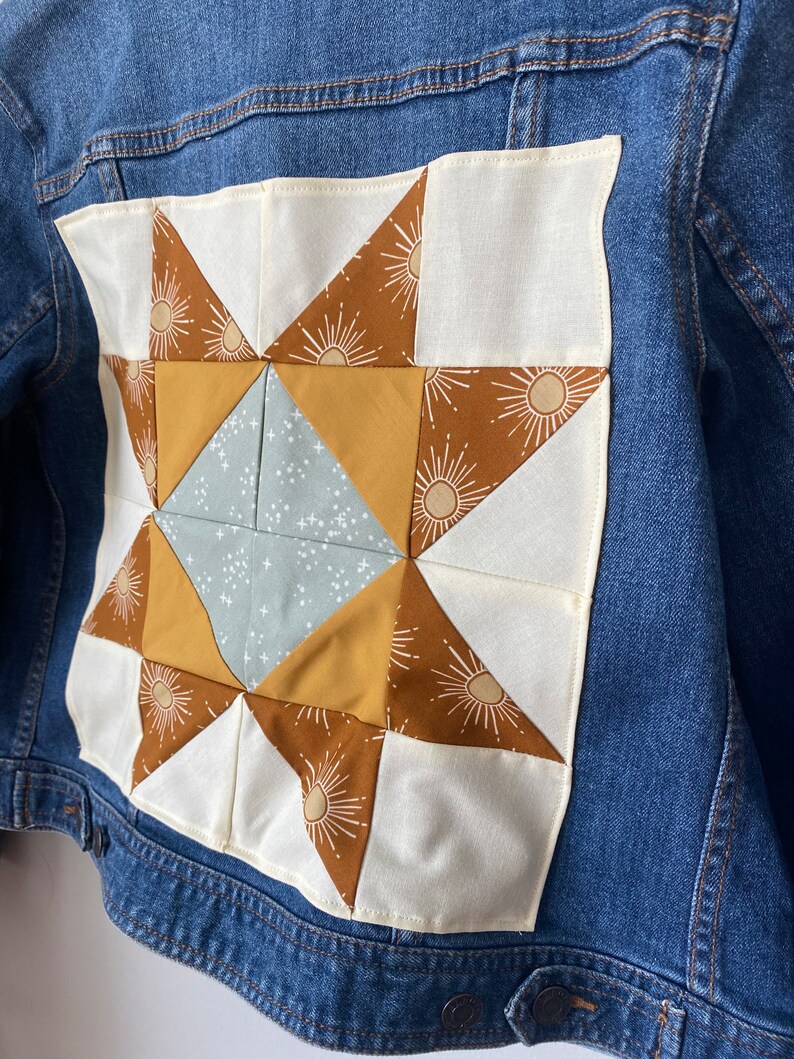May include: A blue denim jacket with a patchwork star design on the back. The star is made of brown, yellow, and blue fabrics, set against a white background. The star design features a sunburst pattern.