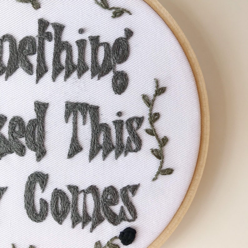 May include: Embroidered hoop art with the text "Something good is coming" in gray thread. The white fabric is accented with green embroidered leaves and a black flower. The wooden hoop frames the artwork.