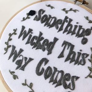 May include: Embroidered hoop art with the phrase "Something Wicked This Way Comes" in gray thread. The white fabric is surrounded by a wooden hoop and features embroidered leaves and a black rose.