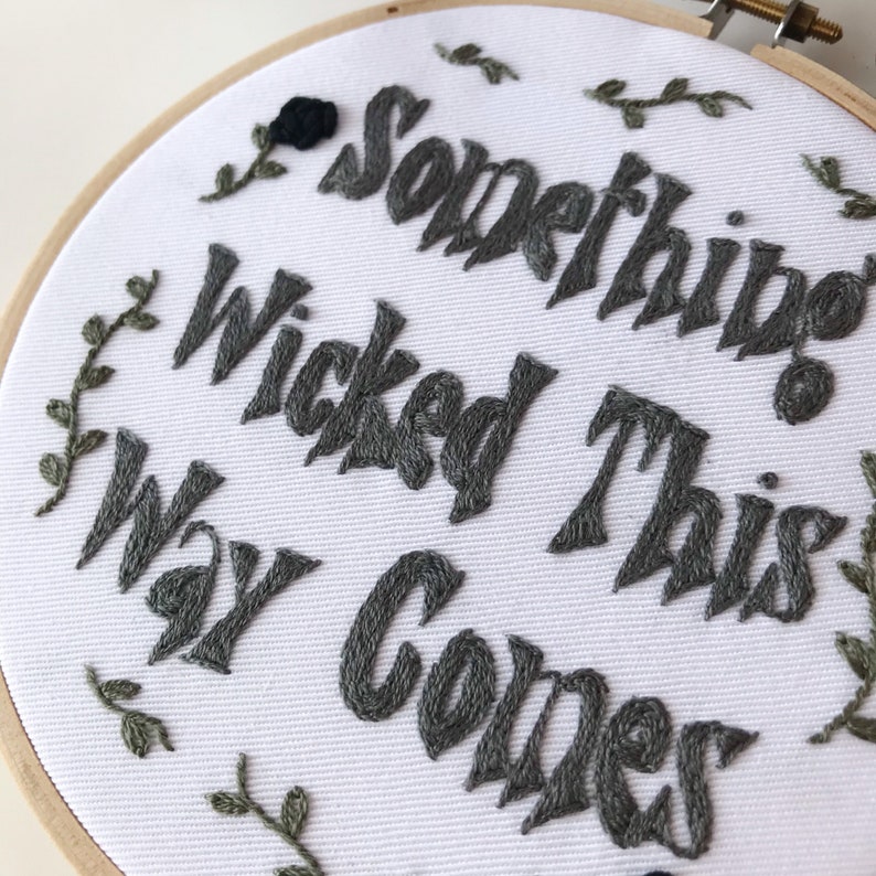 May include: Embroidered hoop art with the phrase "Something Wicked This Way Comes" in gray thread. The white fabric is surrounded by a wooden hoop and features embroidered leaves and a black rose.