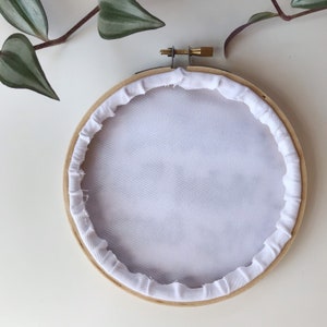 May include: A wooden embroidery hoop with white fabric stretched inside. The fabric is secured with a white, ruffled trim. A brass screw and metal hardware are visible at the top. Green and purple plant leaves are in the background.
