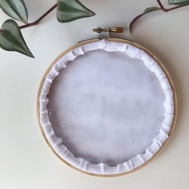 May include: A wooden embroidery hoop with white fabric stretched inside. The fabric is secured with a white, ruffled trim. A brass screw and metal hardware are visible at the top. Green and purple plant leaves are in the background.
