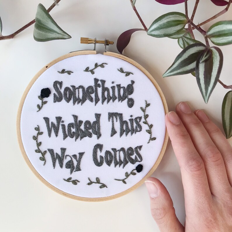May include: A round embroidery hoop with the phrase "Something Wicked This Way Comes" stitched in gray thread on white fabric. The design includes floral accents and is framed by a wooden hoop.