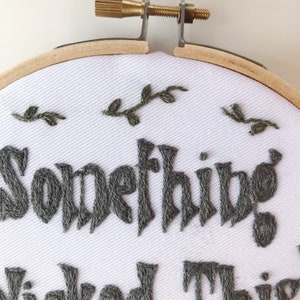 May include: Embroidered hoop art with the words "Something Wicked This Way Comes" in dark gray thread. The white fabric is adorned with small embroidered green leaves, set within a wooden hoop.