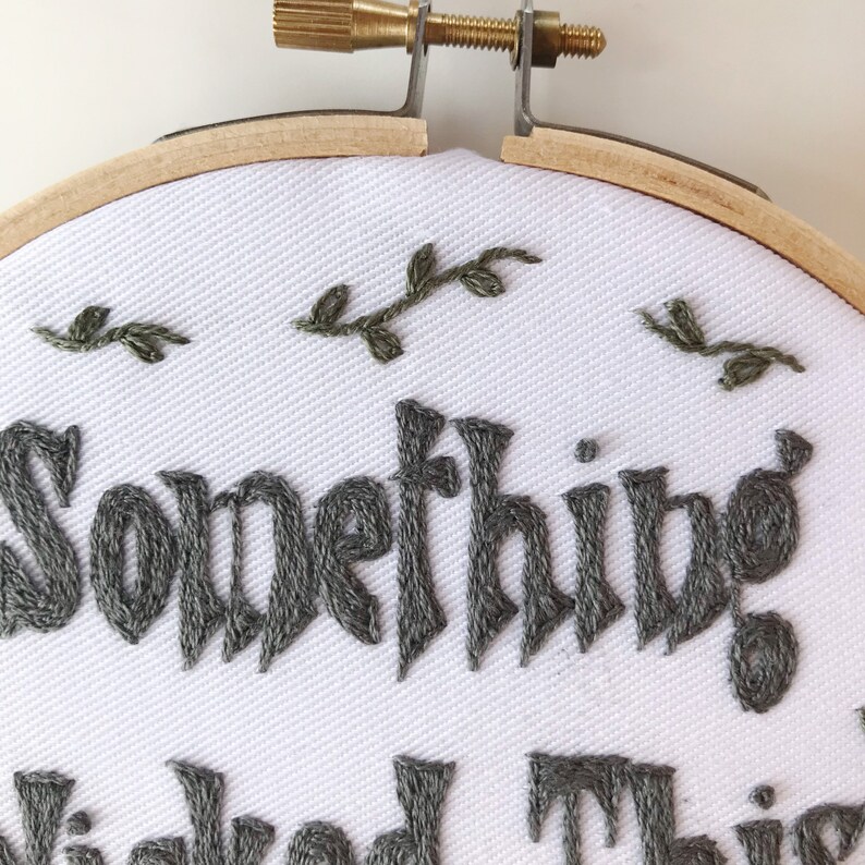 May include: Embroidered hoop art with the words "Something Wicked This Way Comes" in dark gray thread. The white fabric is adorned with small embroidered green leaves, set within a wooden hoop.