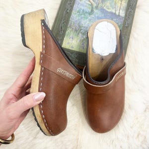 Peut inclure: Une paire de sabots en cuir marron avec des semelles en bois. Les sabots ont une sangle avec le mot "GUSTAVSON" imprimé dessus. Les sabots sont présentés sur une surface blanche et duveteuse.