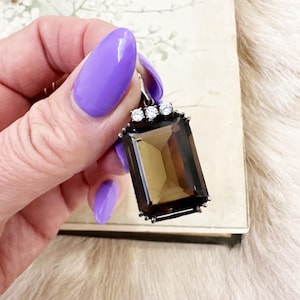 May include: A brown gemstone pendant with a silver setting and four small clear stones. The pendant is held in a hand with purple nail polish.