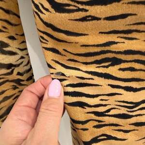 May include: Close-up of fabric with a tiger stripe pattern. The fabric has a tan background with black stripes. The fabric is slightly wrinkled. A hand with pink nail polish is touching the fabric.