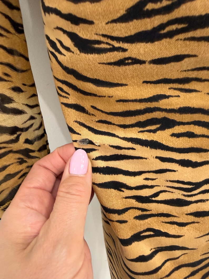 May include: Close-up of fabric with a tiger stripe pattern. The fabric has a tan background with black stripes. The fabric is slightly wrinkled. A hand with pink nail polish is touching the fabric.