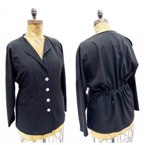 May include: A black, long-sleeved jacket with a button-up front and a drawstring waist. The jacket has a relaxed fit and a collar.