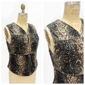 May include: A sleeveless waistcoat featuring a black and tan snakeskin pattern with a full-length front zip closure. The garment has a V-neckline and a structured fit, displaying a textured surface throughout the entire piece on a mannequin.