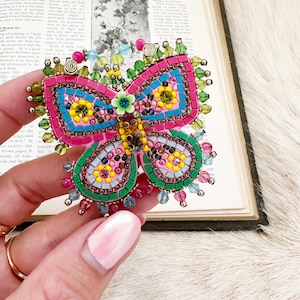 Könnte beinhalten: Eine farbenfrohe Schmetterlingsbrosche mit Mosaikmuster. Die Brosche hat rosa, blaue, grüne und gelbe Akzente mit Perlendetails. Der Schmetterling wird in einer Hand gehalten, mit einem Vintage-Buch im Hintergrund.