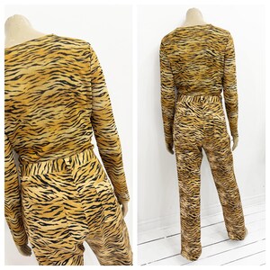 May include: A two-piece outfit featuring a long-sleeved top and matching pants. The ensemble is covered in a tiger stripe pattern, with shades of tan and black. The top has a crew neck, and the pants have a straight leg.