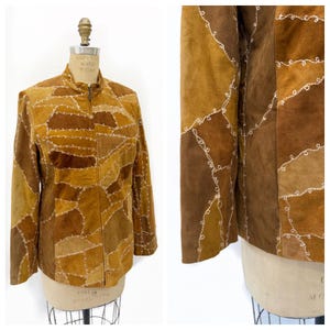 May include: A brown suede jacket with a patchwork design. The jacket has a stand-up collar, a full-length zip, and long sleeves. The seams are accented with white stitching. The jacket is displayed on a mannequin.