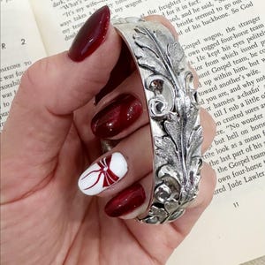 May include: A silver cuff bracelet with an elaborate leaf pattern. The wide bracelet displays detailed carvings of leaves and swirling motifs. The image shows the bracelet being held, emphasising its texture and design.