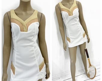 May include: White tennis dress with a V-neckline and tan and peach accents. The sleeveless dress features a fitted silhouette and pockets with matching trim. A vintage wooden tennis racket rests beside the dress, suggesting a retro sports theme.