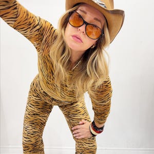 May include: A person wearing a tan and black tiger-striped jumpsuit, brown cowboy hat, and orange-tinted sunglasses. The hat has a decorative silver accent. The person is also wearing multiple bracelets.