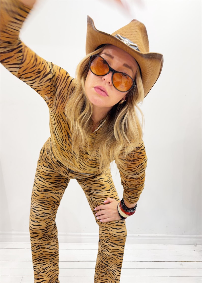 May include: A person wearing a tan and black tiger-striped jumpsuit, brown cowboy hat, and orange-tinted sunglasses. The hat has a decorative silver accent. The person is also wearing multiple bracelets.