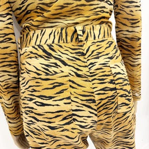 May include: A two-piece outfit featuring a long-sleeved top and high-waisted pants, both with a bold tiger stripe pattern in shades of yellow and black. The pants have back pockets and a fitted waist.