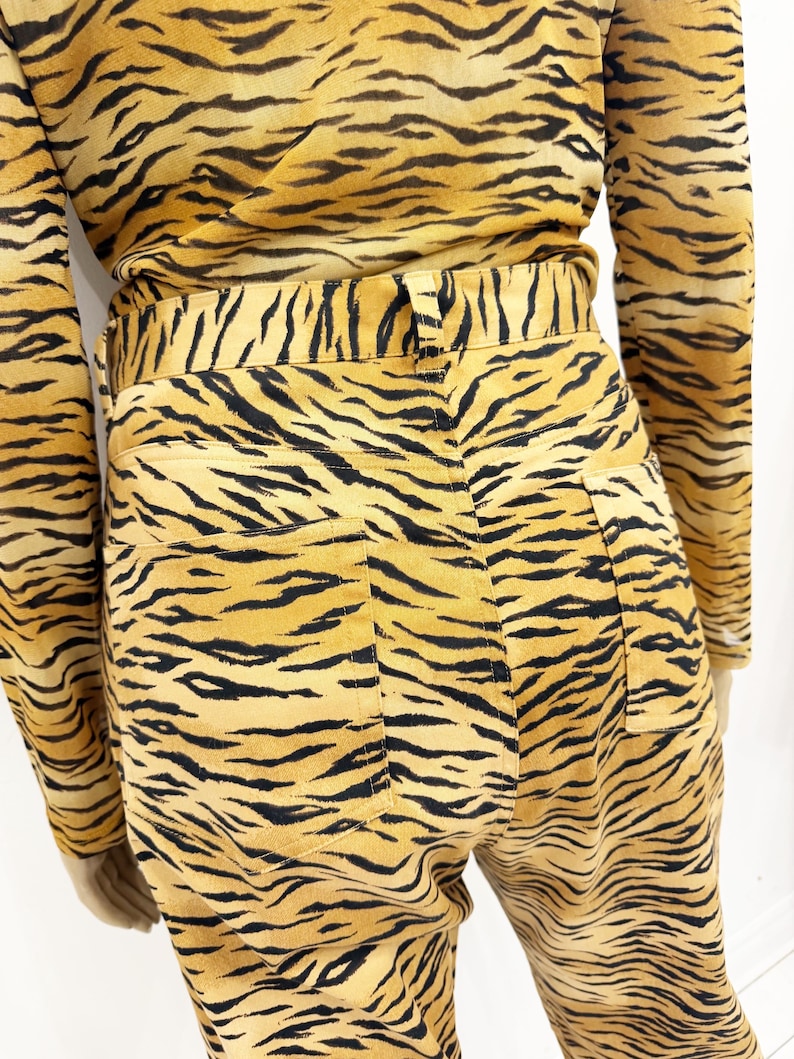 May include: A two-piece outfit featuring a long-sleeved top and high-waisted pants, both with a bold tiger stripe pattern in shades of yellow and black. The pants have back pockets and a fitted waist.