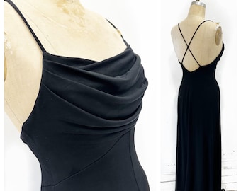 May include: A black evening gown with a cowl neckline and spaghetti straps. The dress has a floor-length skirt and a low back with crisscross straps. The fabric appears to be a smooth, flowing material.