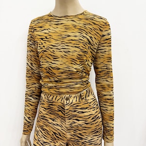 May include: A two-piece outfit featuring a long-sleeved top and matching pants, both with a tiger stripe pattern in shades of tan and black. The top has a crew neck and the pants have a button and zipper closure.