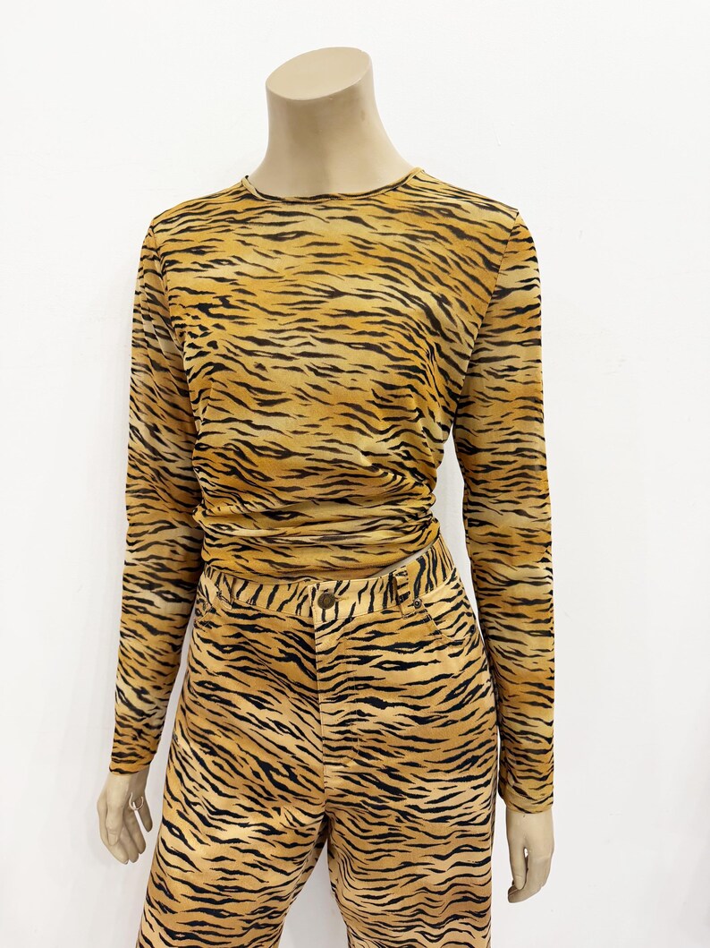 May include: A two-piece outfit featuring a long-sleeved top and matching pants, both with a tiger stripe pattern in shades of tan and black. The top has a crew neck and the pants have a button and zipper closure.