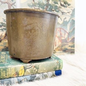 May include: A brass planter with a decorative etched design of a tree and deer. The planter is sitting on top of two books.