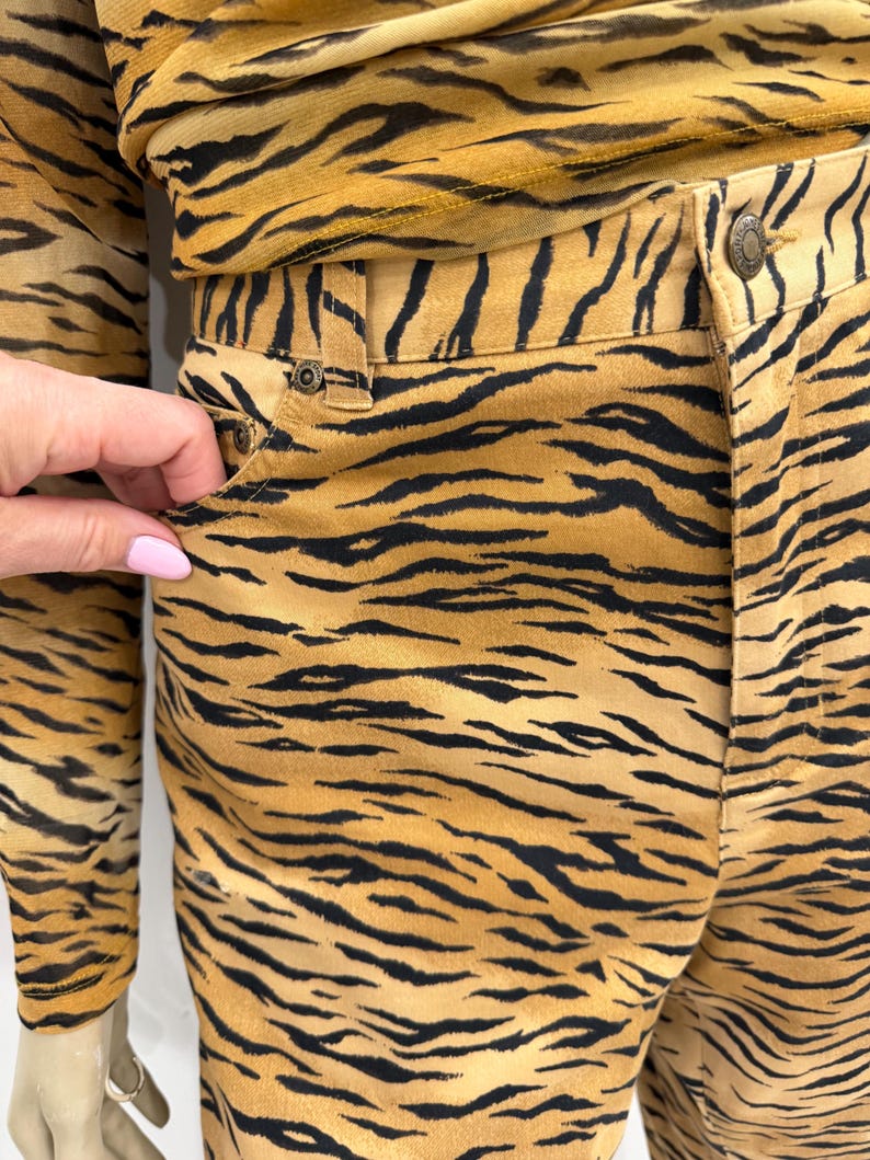 May include: A two-piece outfit featuring a tiger stripe pattern in shades of tan and black. The outfit includes a long-sleeved top and pants. The pants have a button and pocket detail. The fabric appears to be a soft, lightweight material.
