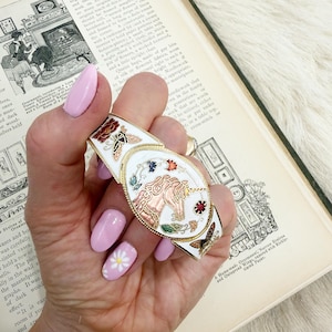 May include: A white and gold hinged bracelet with a central oval design featuring a carousel horse. The bracelet is decorated with colourful floral and butterfly accents. A vintage accessory.