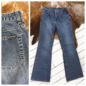 May include: Blue denim jeans with a flared leg. The jeans feature a classic five-pocket design, a button and zip fastening, and a small "MANAGER" label on the back pocket. The jeans are displayed on a white wooden surface.