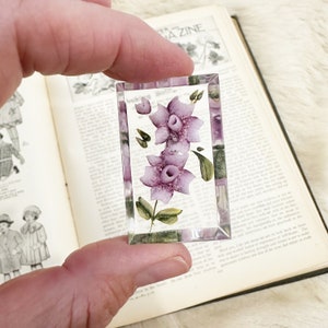 May include: A clear glass paperweight with a hand-painted design of purple flowers with green leaves.