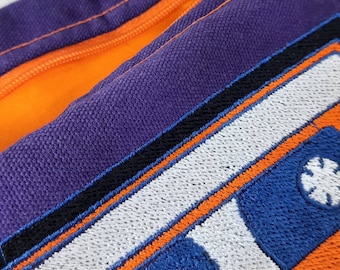 May include: A purple canvas bag with an orange zipper and a large embroidered cassette tape design. The cassette tape is orange, blue, and white.