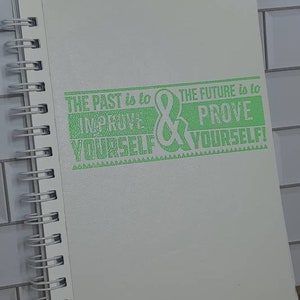 Op de afbeelding: Een wit notitieboek met spiraalbinding en de tekst "The past is to improve yourself & the future is to prove yourself!" in groen.