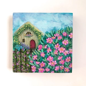 May include: A square painting on wood featuring a small cottage with a red door, a yellow vine-covered roof, and a dark fence. The foreground is filled with vibrant pink flowers and green foliage against a light blue sky with visible brushstrokes.
