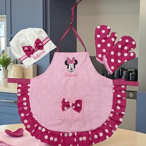 Könnte beinhalten: Eine rosa Schürze mit Minnie-Mouse-Grafik und dem eingestickten Namen Rebecca auf der Vorderseite, mit gepunktetem Rüschensaum, einer mittigen Tasche mit Schleife, einer passenden weißen Kochmütze mit rosa Schleife und rosa gepunkteten Ofenhandschuhen.