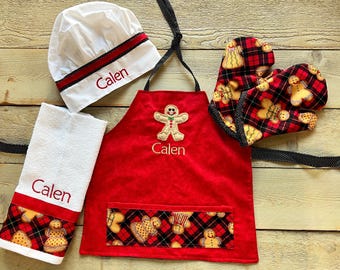 May include: A red gingerbread-themed children's apron with a gingerbread man design and the name "Calen" embroidered on it. The set also includes a white chef's hat with a red band and the name "Calen" embroidered on it, a white kitchen towel with a red gingerbread-themed border and the name "Calen" embroidered on it, and a pair of red and black gingerbread-themed oven mitts.