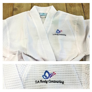 May include: White waffle-weave robe with a shawl collar. The robe features an embroidered butterfly logo in blue, purple, and red, along with the text "SA Body Contouring" in black. The robe is designed for comfort and relaxation.