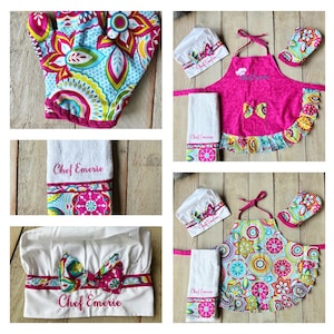 May include: A pink and white chef's hat, a pink apron with a colorful floral pattern, a white towel with the text "Chef Emerie", and a colorful floral oven mitt. The apron has a pink bow and a ruffled hem.