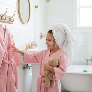 May include: A light pink waffle-weave robe with an embroidered initial hangs on a wooden rack with brass hooks. A young person wearing a matching robe and a towel on their head holds a teddy bear in a bright bathroom setting.